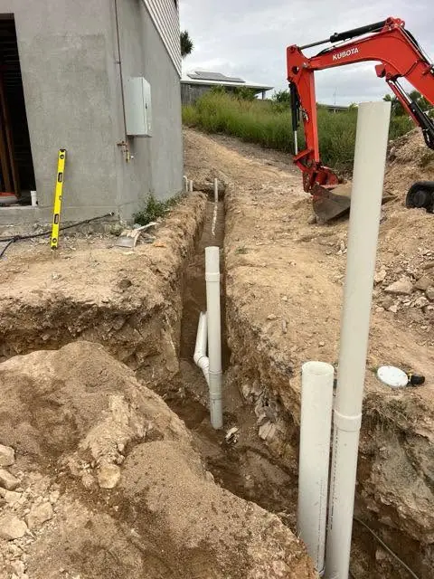 A drain trench with riser pipes beside a new slab, red excavator behind